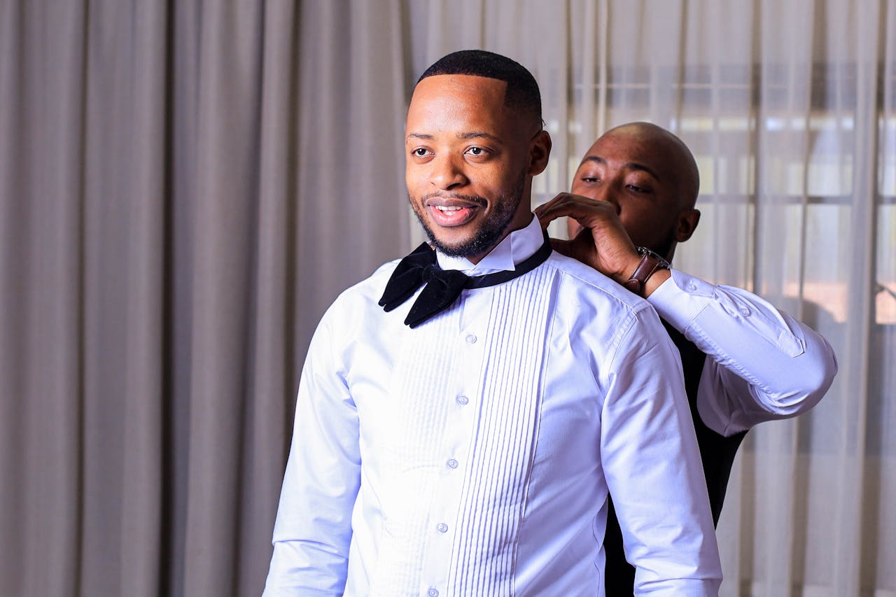 A man helps the groom adjust his bow tie before the wedding ceremony. Happy and elegant moment captured indoors.